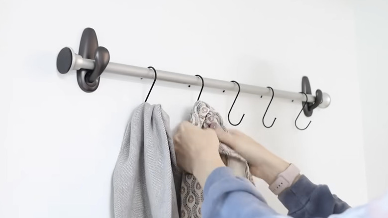 Kadının elleri, ev yapımı raylı rafa iliştirilmiş kancalara eşarp asıyor.