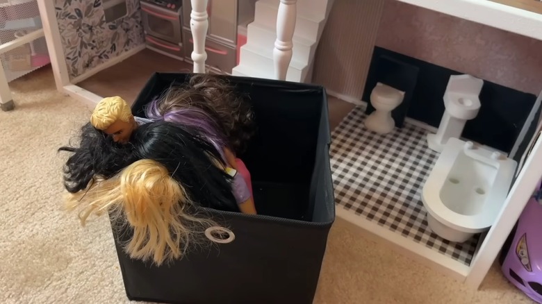 Bir oyuncak bebek evinin yanına yerleştirilmiş Barbie bebekleriyle dolu siyah kumaş saklama küpü.