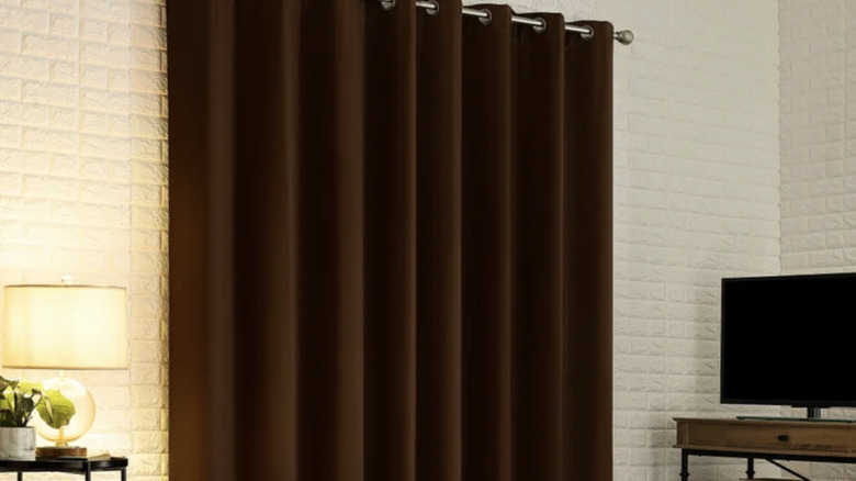 Bir pencerenin üzerinde sergilenen kahverengi Sun Zero Broderick karartma perdelerinin ürün resmi. Oturma odası, sehpa üzerinde TV ve üstünde masa lambası bulunan bir komidin.