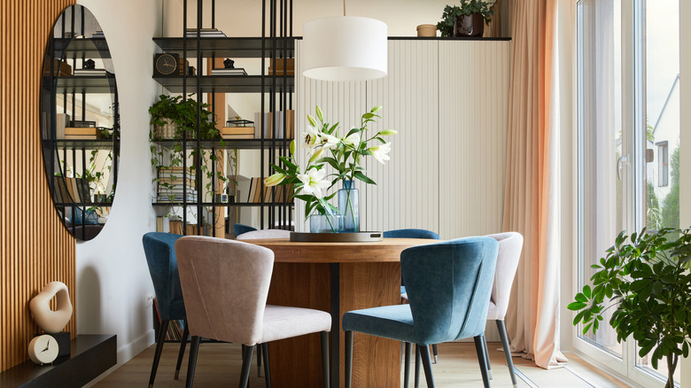 Yuvarlak ahşap masa, mavi ve beyaz döşemeli sandalyeler ve duvar aynası bulunan modern yemek odası.