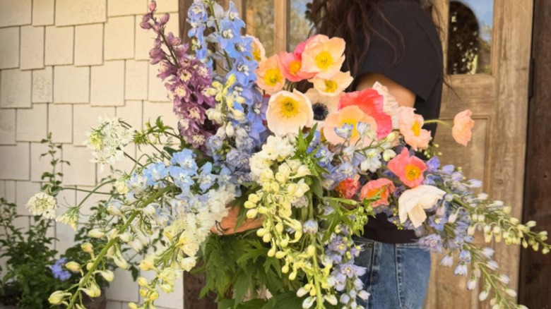 Joanna Gaines verandasında büyük boy bir haşhaş ve delphinium buketi tutuyor.