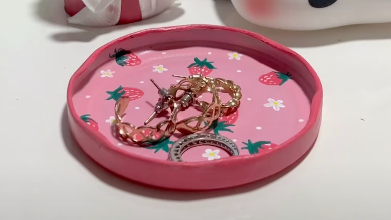 Pembeye boyanmış, çiçekler ve çileklerle süslenmiş, içinde küçük mücevher eşyaları bulunan eski bir kavanoz kapağı.