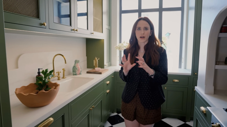 Rachel Brosnahan yeşil bir mutfakta duruyor.