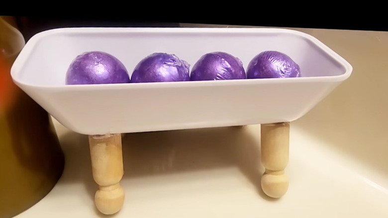 Ahşap mandal bebeklerden yapılmış bir DIY banyo tepsisi ve içinde mor banyo bombaları olan plastik bir tereyağı tabağı.