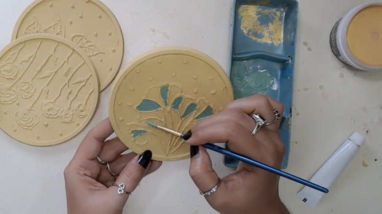 Bir kadın, sıcak tutkal desenleriyle süslenmiş altın renkli plastik kapaklara turkuaz boya sürüyor.