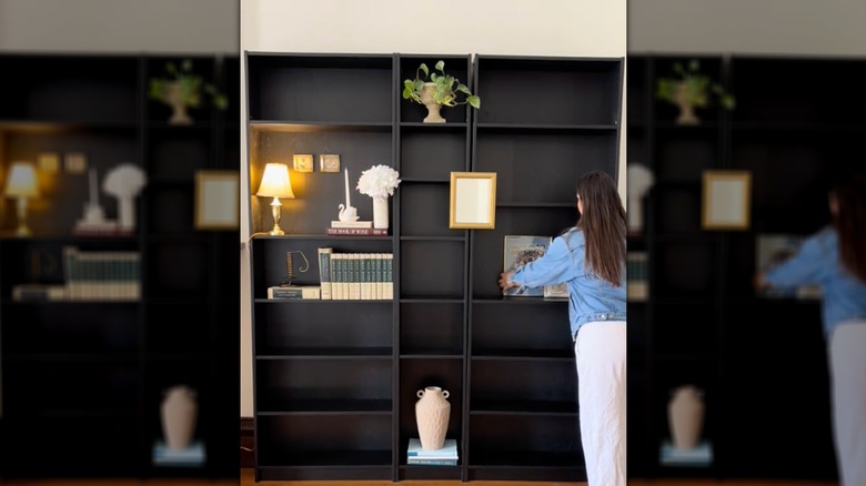 Bir kadın evinin duvarını kaplayan iki büyük siyah kitap rafına süs eşyaları düzenliyor.