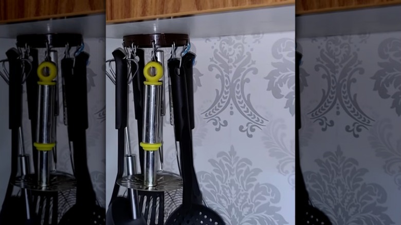 Üst dolabın alt kısmına tutturulmuş bir fıstık ezmesi kavanozunun plastik vidalı kapağına tutturulmuş kancalardan sarkan mutfak eşyaları.
