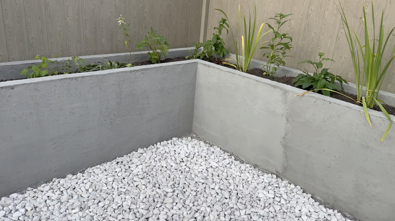 Beyaz çakıllarla kaplı arka bahçenin çitlerle çevrili köşesinde bitkilerle dolu beton bir çiçeklik yer alıyor.