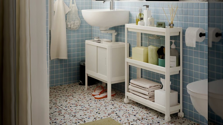 Mavi fayans duvarın önünde tekerlekli beyaz bir banyo malzeme arabasını gösteren IKEA katalog fotoğrafı