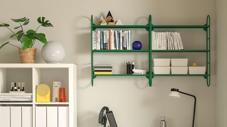 Duvara monte edilen yeşil IKEA duvar rafında kitaplar, kırtasiye malzemeleri ve süs eşyaları bulunur.