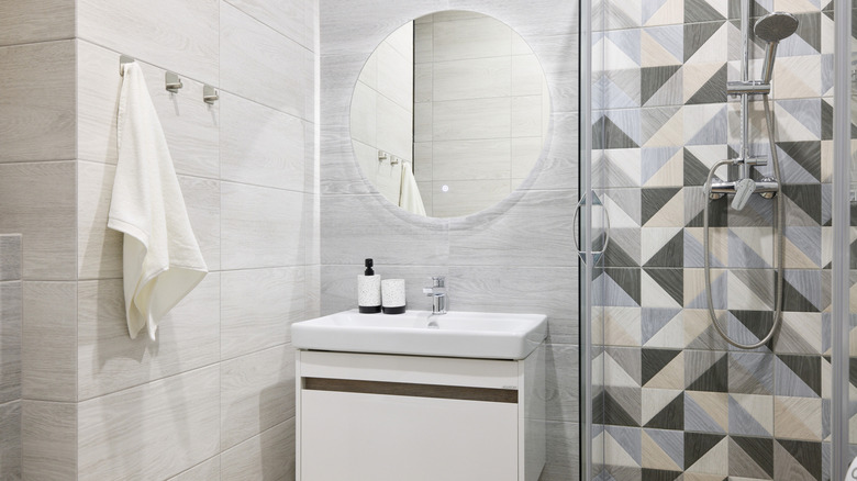 Lavabo ve makyaj ünitesinin yanında siyah, gri ve krem ​​renkte geometrik döşemelere sahip bir duş ve üstündeki duvarda büyük bir dairesel ayna yer alıyor.