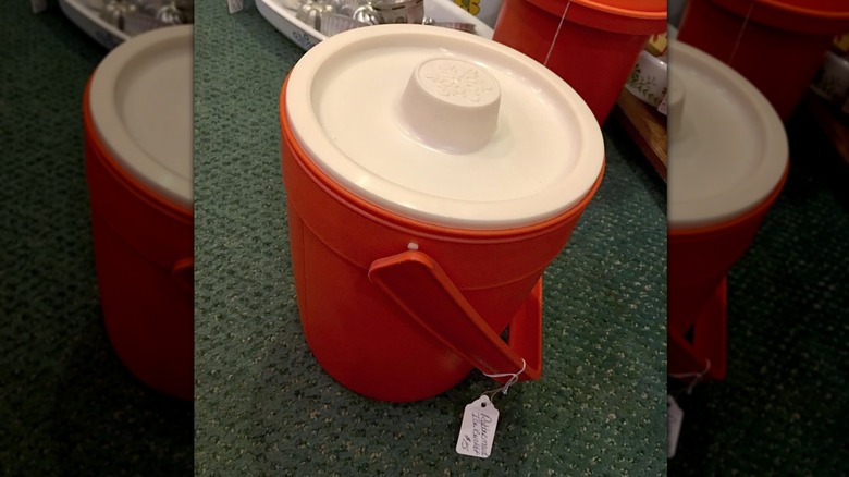 Yeşil halının üzerinde duran turuncu ve beyaz vintage bir Rubbermaid buz kovası.