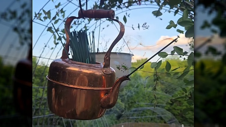 Üst kısmında sitronella çubuklarını tutan bir kancaya asılı bakır bir su ısıtıcısı ve ağzında yanan bir çubuk var.