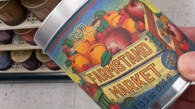 Bir el, bir mağazanın koridorunda Hobi Lobisinden alınan Farmstand Market teneke mumunu tutuyor.