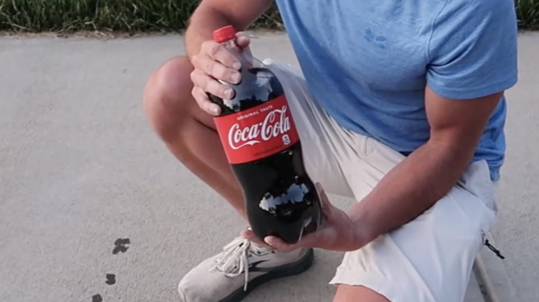 Bir adam beton bir garaj yolunda çömelip elinde bir şişe Coca-cola tutuyor.