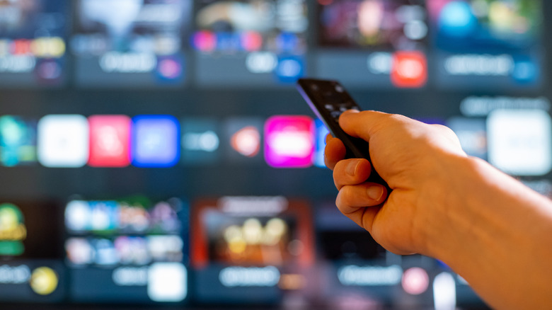 Uzaktan kumandayı tutan bir el, onu arka planda bulanık uygulamalar bulunan bir TV'ye doğrultuyor