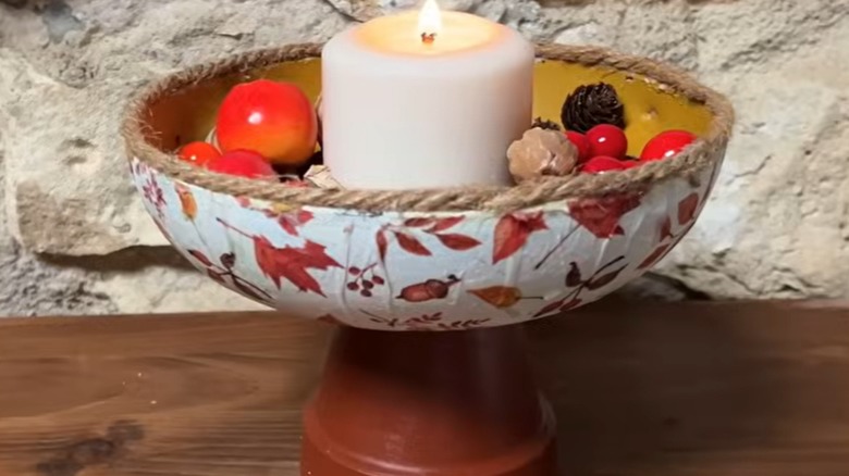 Eski bir kızartma tavasından yapılmış bir kapta bir mum ve dekoratif suni meyve bulunur.