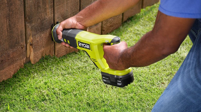 Altında çimen bulunan ahşap bir çit üzerinde Ryobi panter testere kullanan kişi