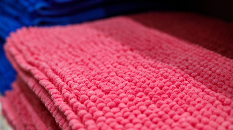 Arka planda birkaç mavi havlu bulunan katlanmış pembe tüylü bir banyo paspası.
