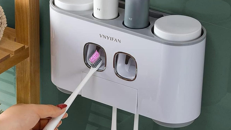 Banyo duvarına yapıştırılmış diş macunu dağıtıcısı ve tutucusu, bir kadın eliyle dağıtıcıya diş fırçasını yerleştiriyor