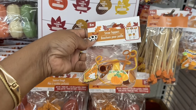 Dollar Tree'de inek baskılı başparmak tırnağı ve altın bilezikleri olan ve sonbahar temalı saman toppers paketi tutan bir kişi