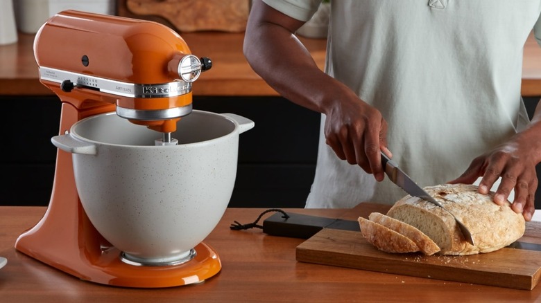 Birisi turuncu KitchenAid stand mikserinin yanında bir somun taze ekmek kesiyor