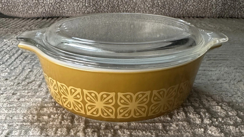 Sonbahar Çiçek deseniyle süslenmiş hardal renginde vintage Pyrex cam kapaklı güveç tabağı, kumaş bir kanepenin üzerinde duruyor.