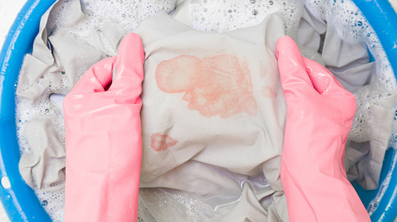 Pembe eldivenli bir kişi kovada kırmızı lekeli kıyafetleri yıkıyor