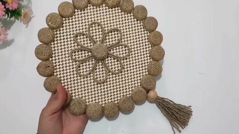 Bir el, çuval, kaymaz paspas, sicim, püskül ve ahşap boncuktan yapılmış kendin yap asılı duvar resimlerini tutuyor.