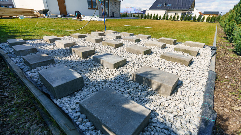Arka bahçe kulübesi için peyzaj kumaşı, çakıl ve beton bloklardan oluşan bir temel.