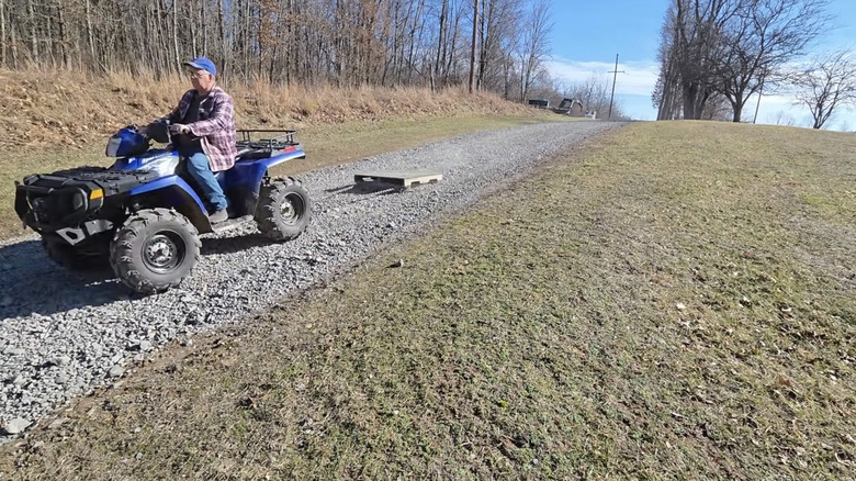 Mavi şapkalı ve flanel gömlekli bir adam, mavi bir ATV'ye binerek çakıllı bir garaj yolunda ahşap bir paleti sürüklüyor.