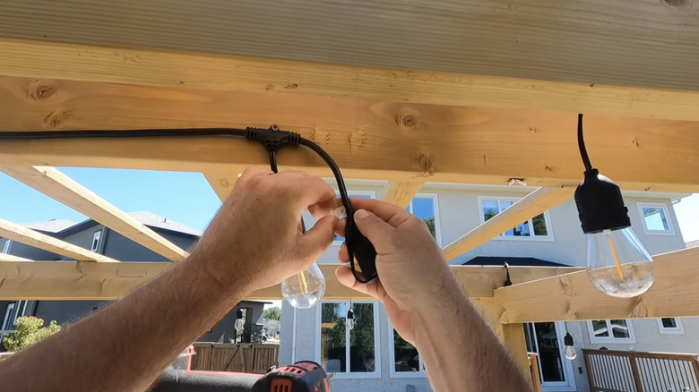 Bir kişi ahşap bir pergola çatısına büyük Edison ampulleriyle ip ışıkları asıyor.