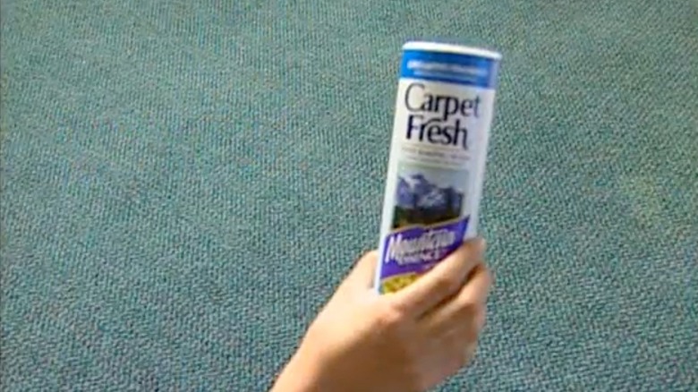 Bir kişinin elinde mavi halının üzerinde tutulan bir kutu Carpet Fresh.