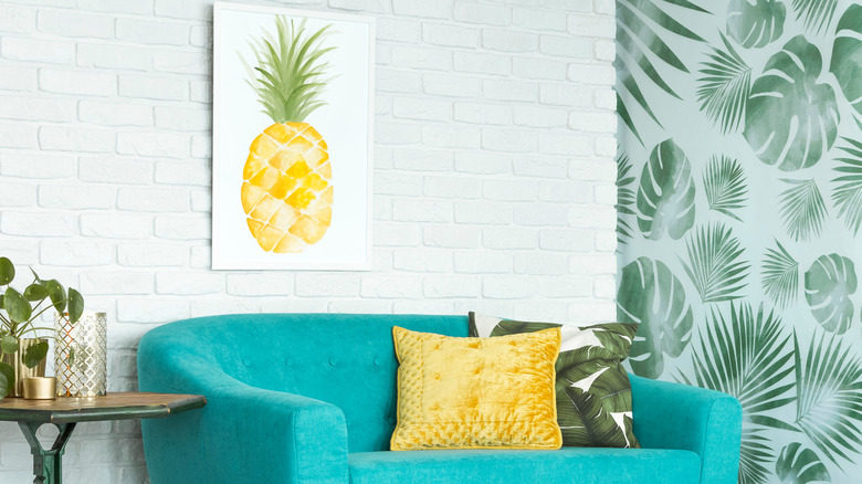 Yaprak desenli duvar kağıdının yanında deniz mavisi kanepe ve çerçeveli ananas sanat resmi bulunan bir oda.
