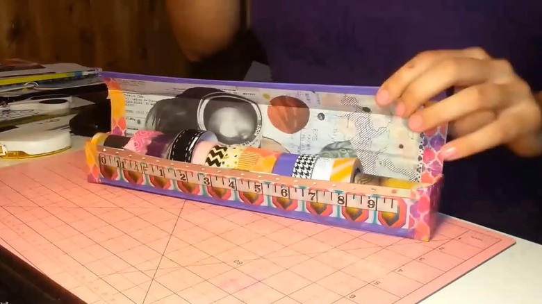 Bir masanın üzerinde DIY washi bant dağıtıcı kutusunu açık tutan bir el