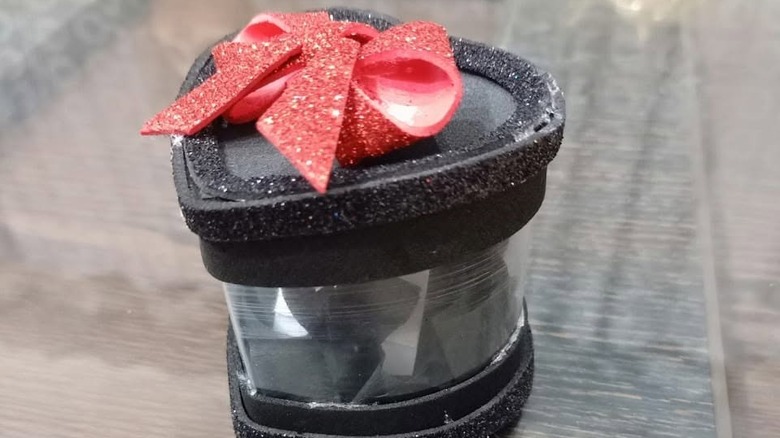 Plastik şişeden yapılmış, kapağının üstünde kırmızı parıltılı fiyonk bulunan, ışıltılı siyah kalp şeklinde biblo kutusu.