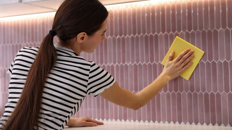 Bir kadın porselen fayanslı mutfağın arka yüzeyini kuru sarı mikrofiber bir bezle siliyor.