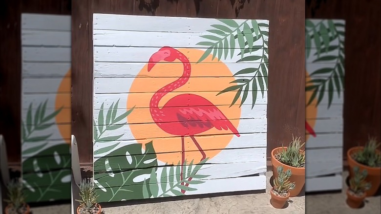Eski bir ahşap palet, üzerine flamingo, güneş ve yeşil bitkilerin boyandığı bir tabloya dönüştürüldü.